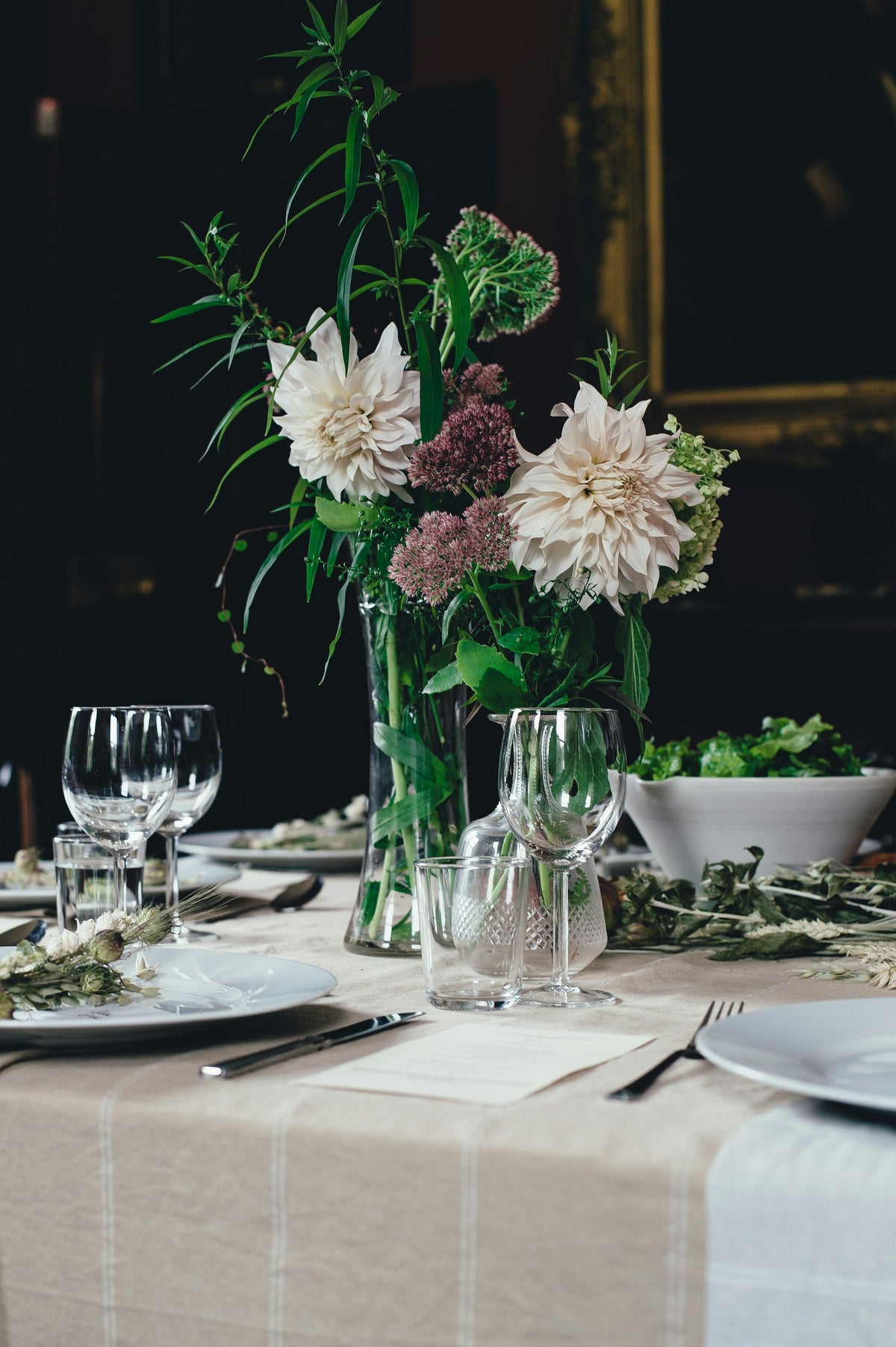 A table set for dinner on the linen tablecloth, with glassware, plates and a vase of dahlias.