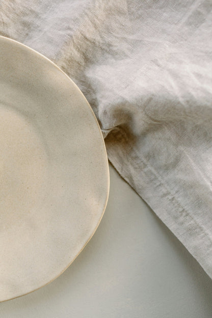 The Linen Tablecloth in undyed washed flax, laid flat beside a cream stoneware plate.