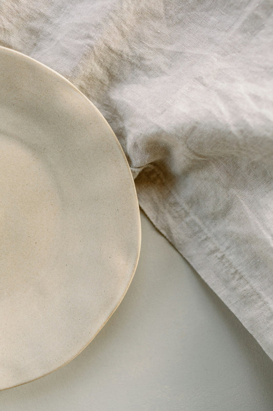 The Linen Tablecloth in undyed washed flax, laid flat beside a cream stoneware plate.